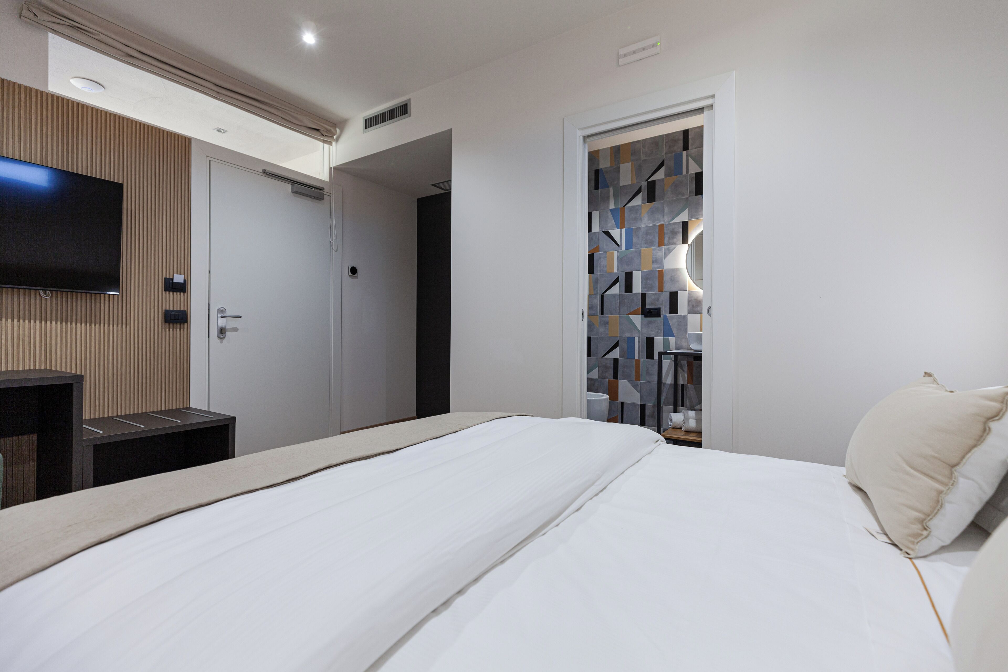 A modern hotel room with a large bed, a flat-screen TV mounted on the wall, a wooden panel wall, and a door with a geometric tile design.