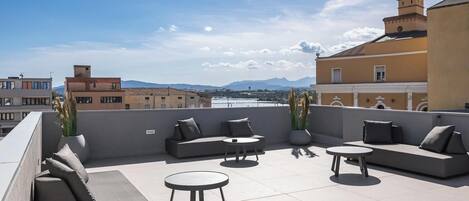 A rooftop terrace with modern outdoor seating, a view of the cityscape, and a body of water in the distance.