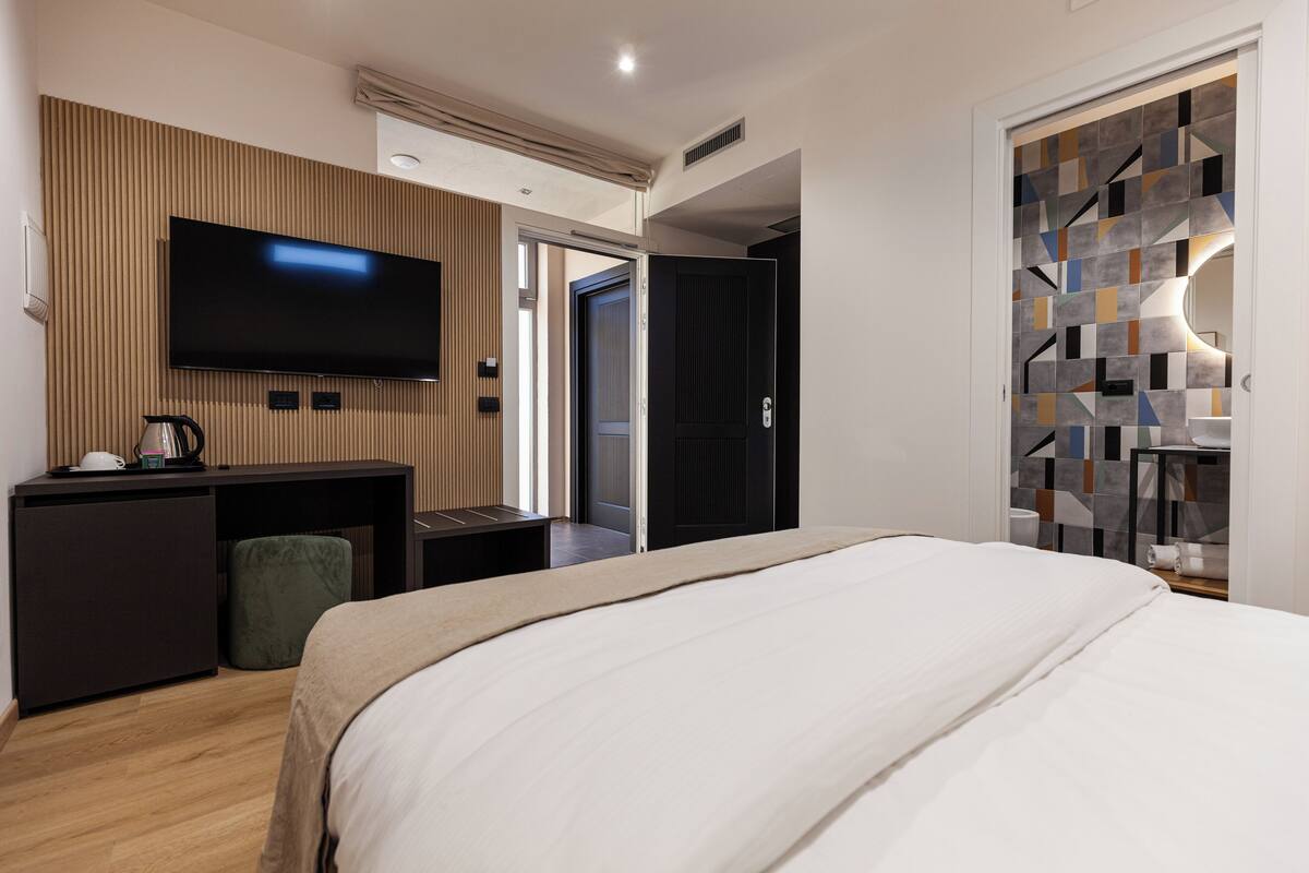 Une chambre d’hôtel moderne dotée d’un grand lit, d’un téléviseur à écran plat fixé au mur, d’un bureau avec une chaise et d’une armoire dont la porte est ornée d’un motif géométrique.