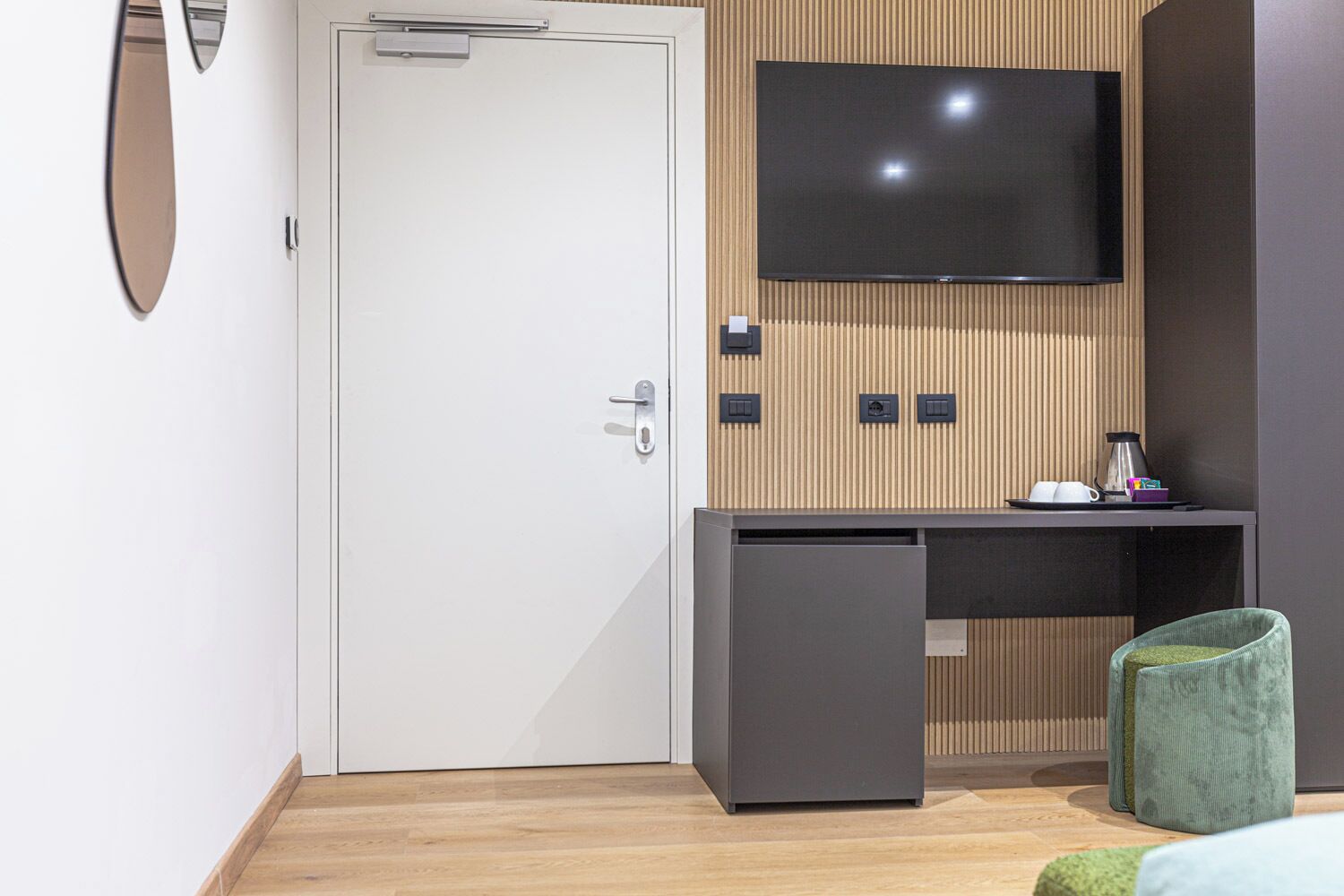 A modern hotel room with a white door, a small desk with a computer, a flat-screen TV mounted on a wooden panel, and a green chair.