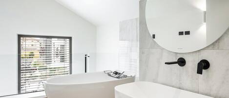 Une salle de bain moderne avec deux baignoires blanches sur pieds, un grand miroir et une fenêtre avec des stores.