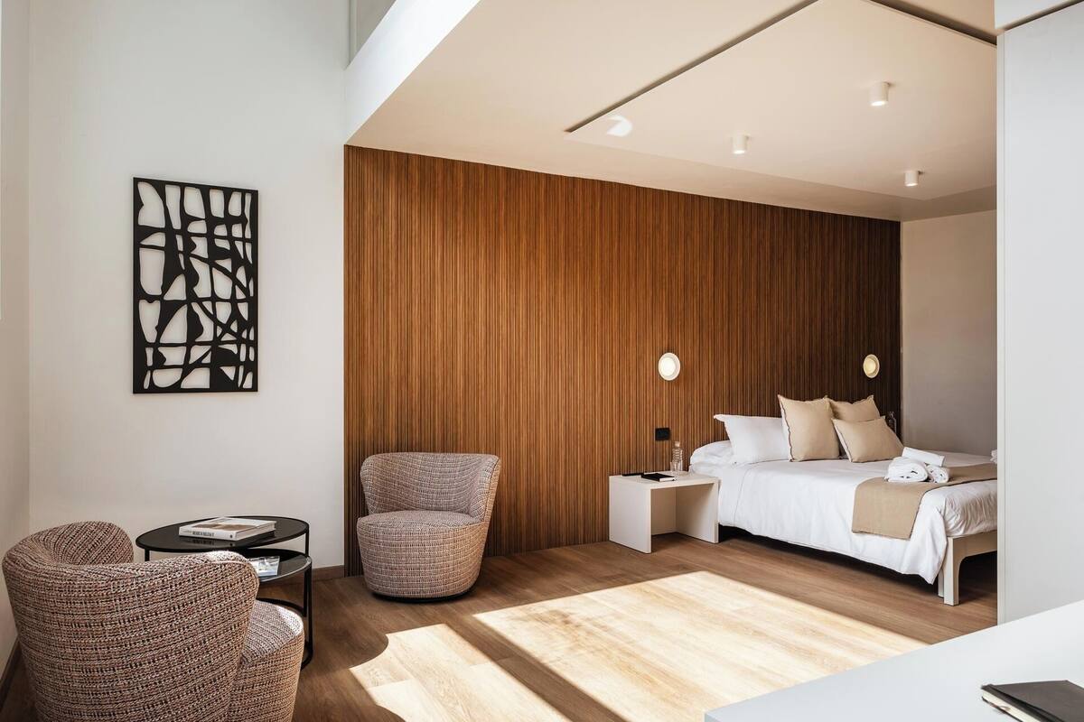 Une chambre d’hôtel moderne avec un mur d’accent en bois, un lit avec du linge de maison blanc, une chaise ronde en osier, une petite table d’appoint et un luminaire fixé au mur.