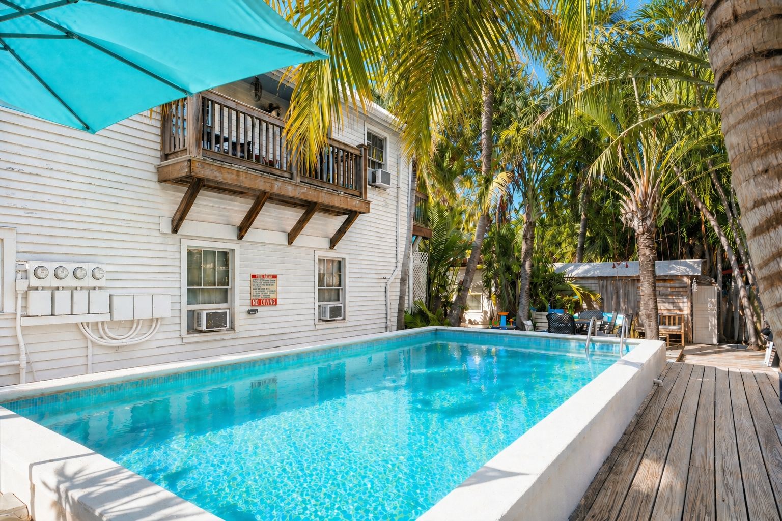 Ein Schwimmbecken mit Holzterrasse, ein Balkon mit Geländer und Palmen.