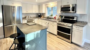 A modern kitchen with white cabinets, stainless steel appliances, a granite countertop, and a central island with bar stools. - 6 Richards Way (Sandwich)