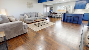 A modern living room with a sofa, coffee table, and a kitchen area with blue cabinets and a bar stool. - 12 Minutes from Crossbar and Turner Falls, Sleeps up to 10, Fast Wi-Fi (Davis)