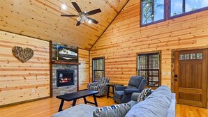 A cozy cabin interior with a fireplace, wooden walls, a ceiling fan, and a large window.