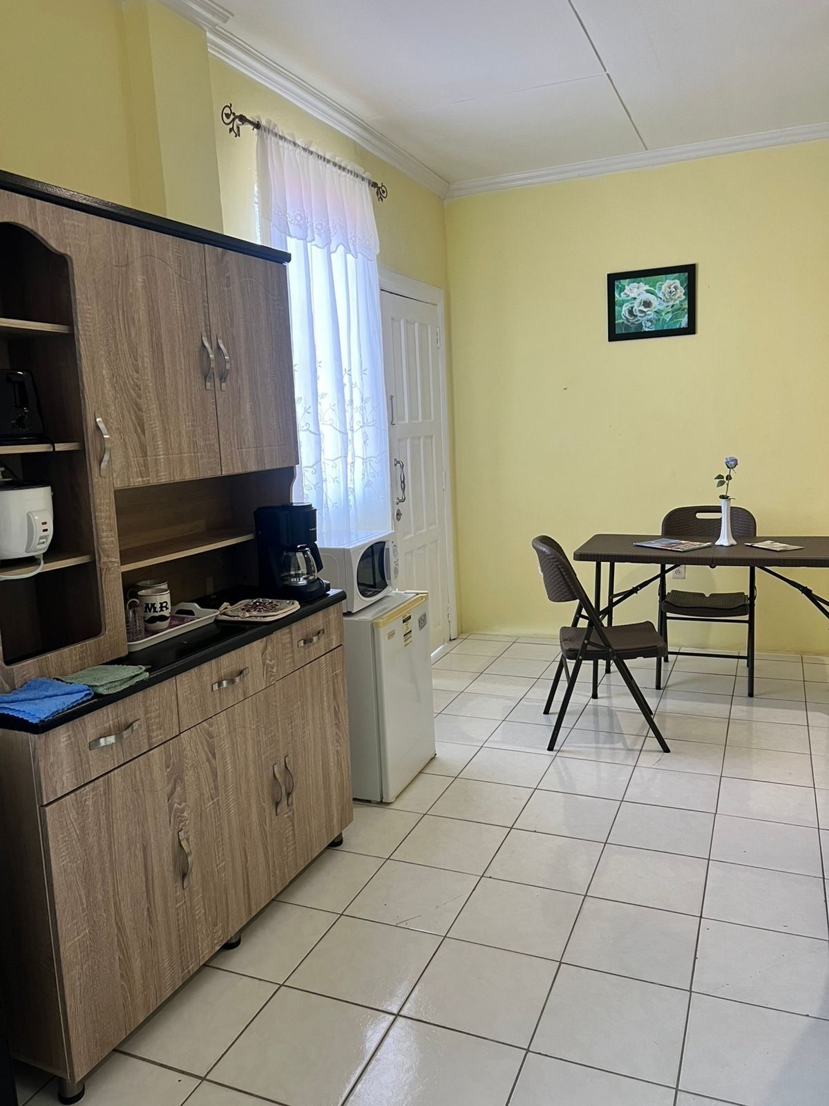 A compact kitchen with a wooden cabinet, a microwave, and a refrigerator. There is a dining table with chairs and a small vase with a flower.