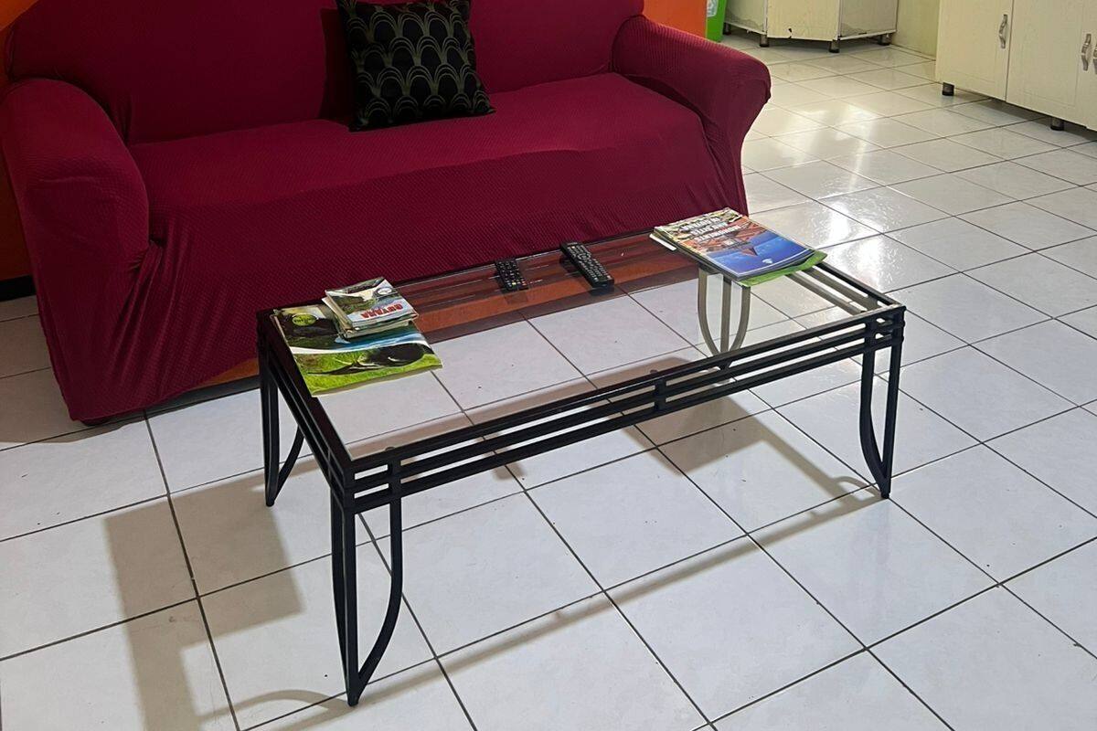 A living room with a red sofa, a glass coffee table with magazines, a kitchen area with cabinets and a refrigerator, and a window with curtains.