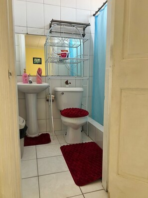 A small bathroom with a white sink, toilet, and shower area. There is a metal drying rack above the toilet and a blue shower curtain. - Emerald Aparment (Georgetown)