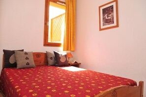 Een slaapkamer met een houten bedframe, een rood bed met bloemenprint en een raam met gele gordijnen.