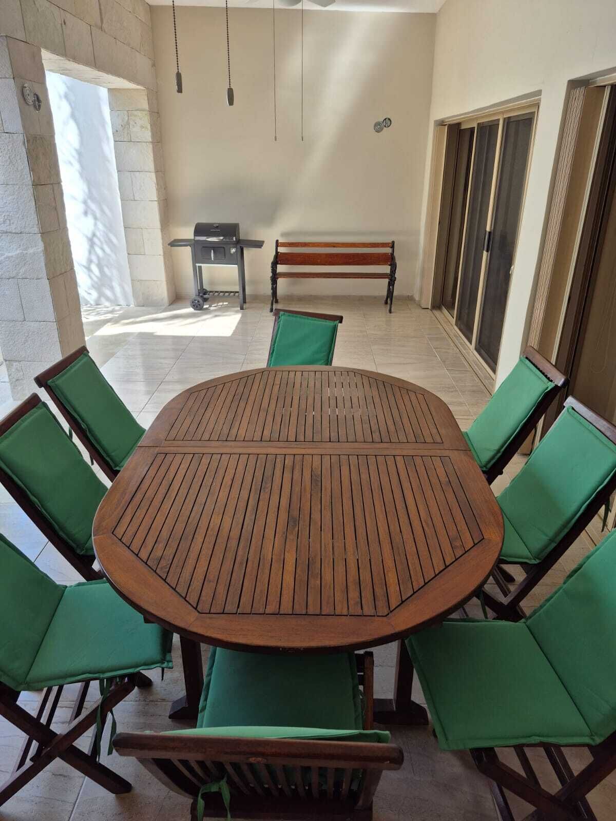 Une table ronde en bois, entourée de six chaises vertes, d’un banc et d’un chariot-grill, dans un patio ensoleillé doté d’un sol carrelé et de murs en pierre.