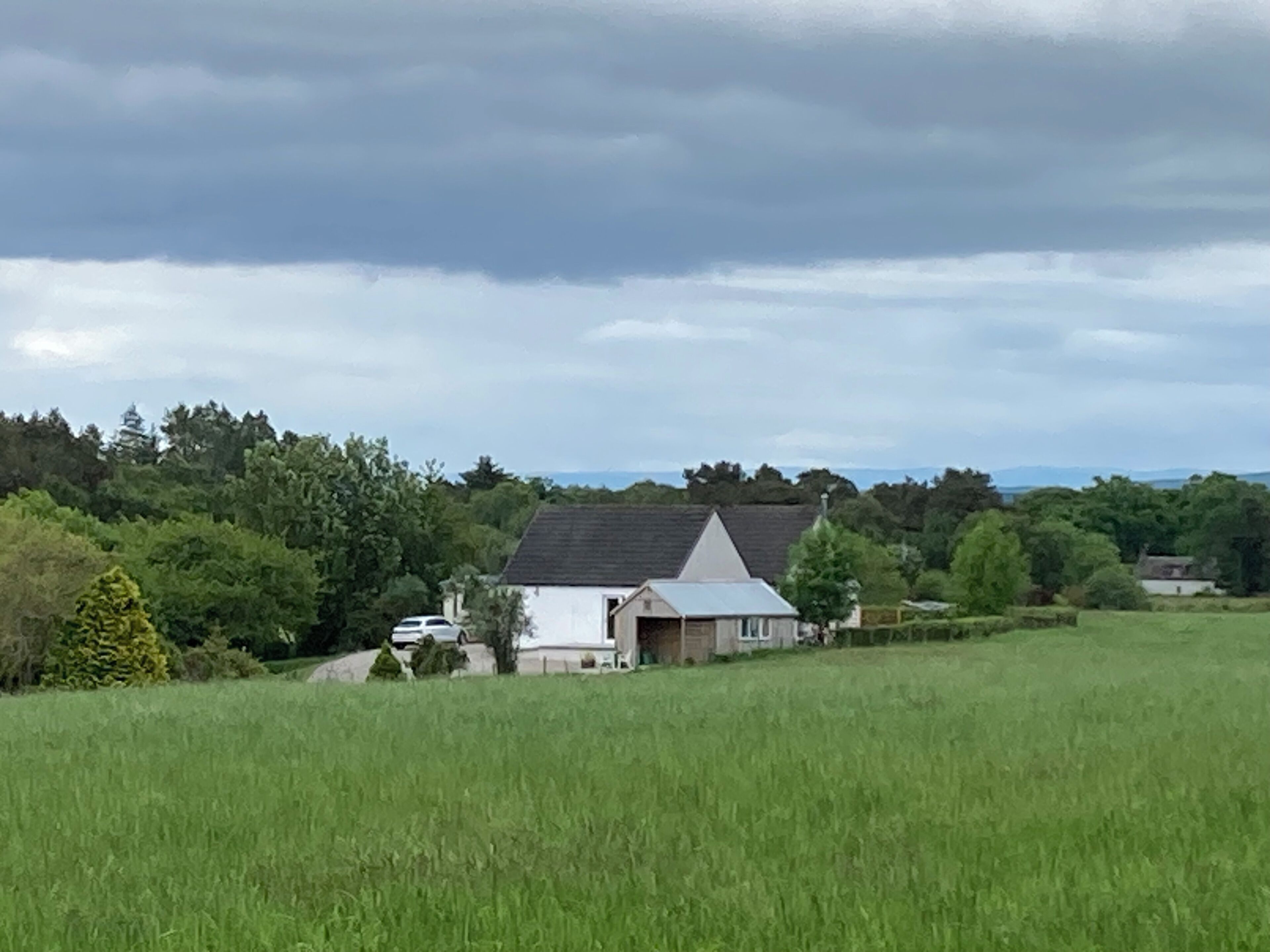Une maison de campagne avec un toit en pente, entourée d’un champ verdoyant et d’une forêt dense.