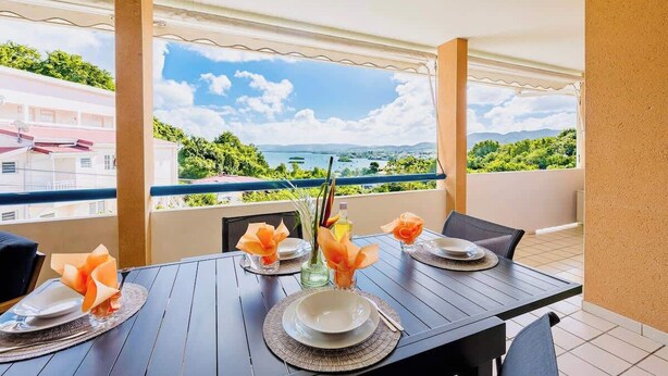 A balcony with a table set for dining, overlooking a scenic landscape with greenery and buildings.