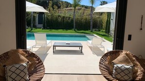 Una habitación con puerta de vidrio que da a una zona de piscina, dos sillas de mimbre con cojines, una mesita y vista a un paisaje verde con árboles y una colina.
