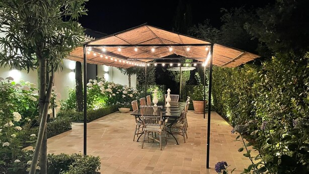 Un patio al aire libre con mesa y sillas cubiertas, rodeado de abundante vegetación y plantas en macetas.