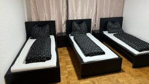 Tres camas individuales con estructura negra y ropa de cama blanca, dispuestas en fila sobre un suelo de madera, con cortinas beige al fondo.