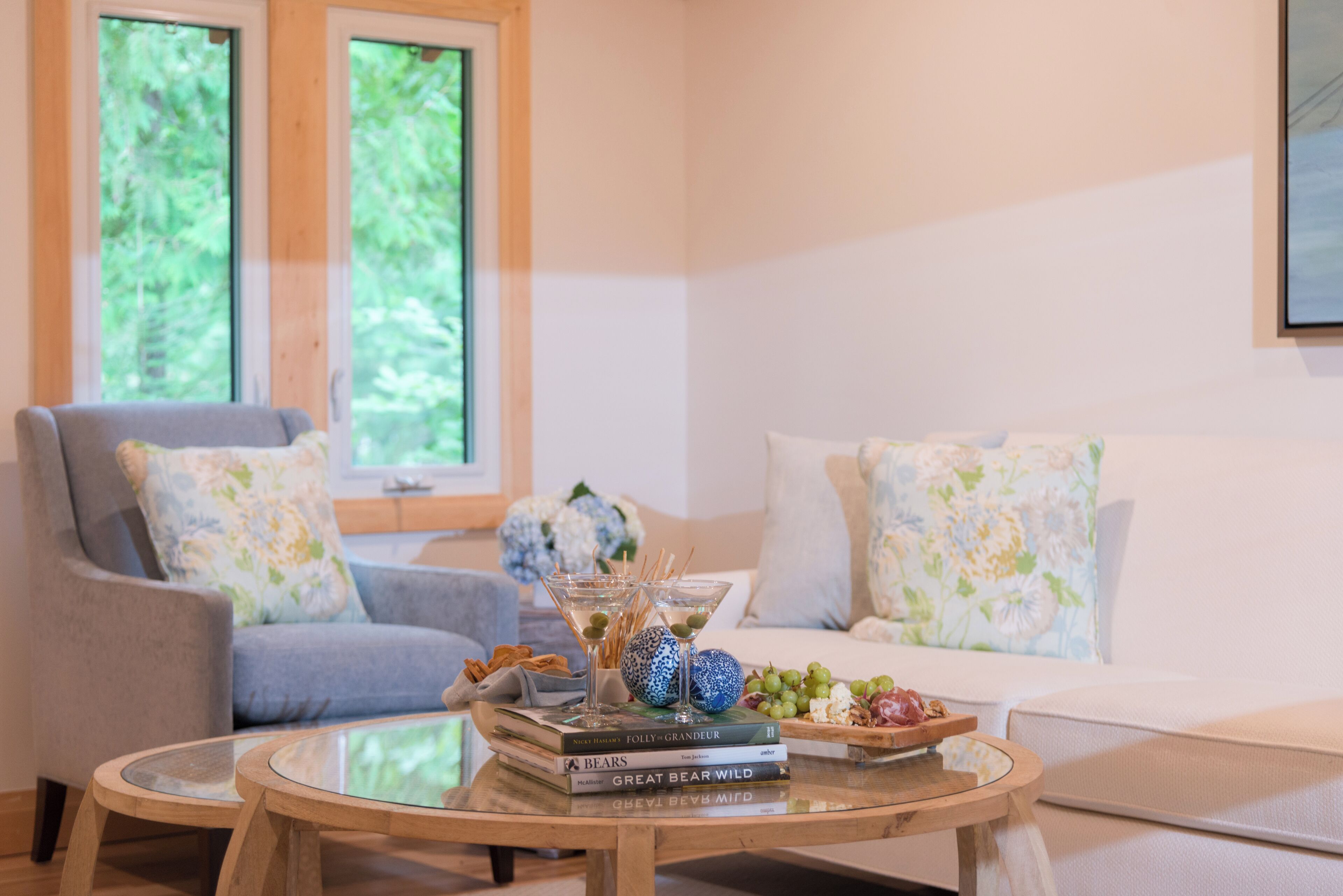 Una sala de estar con un sillón gris, un sofá blanco, una mesa de centro de vidrio y una ventana con vista a follaje.