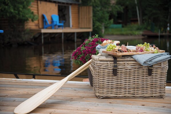 Un panier en osier contenant divers aliments est posé sur une terrasse en bois, près d’une étendue d’eau, avec une pagaie appuyée contre lui.