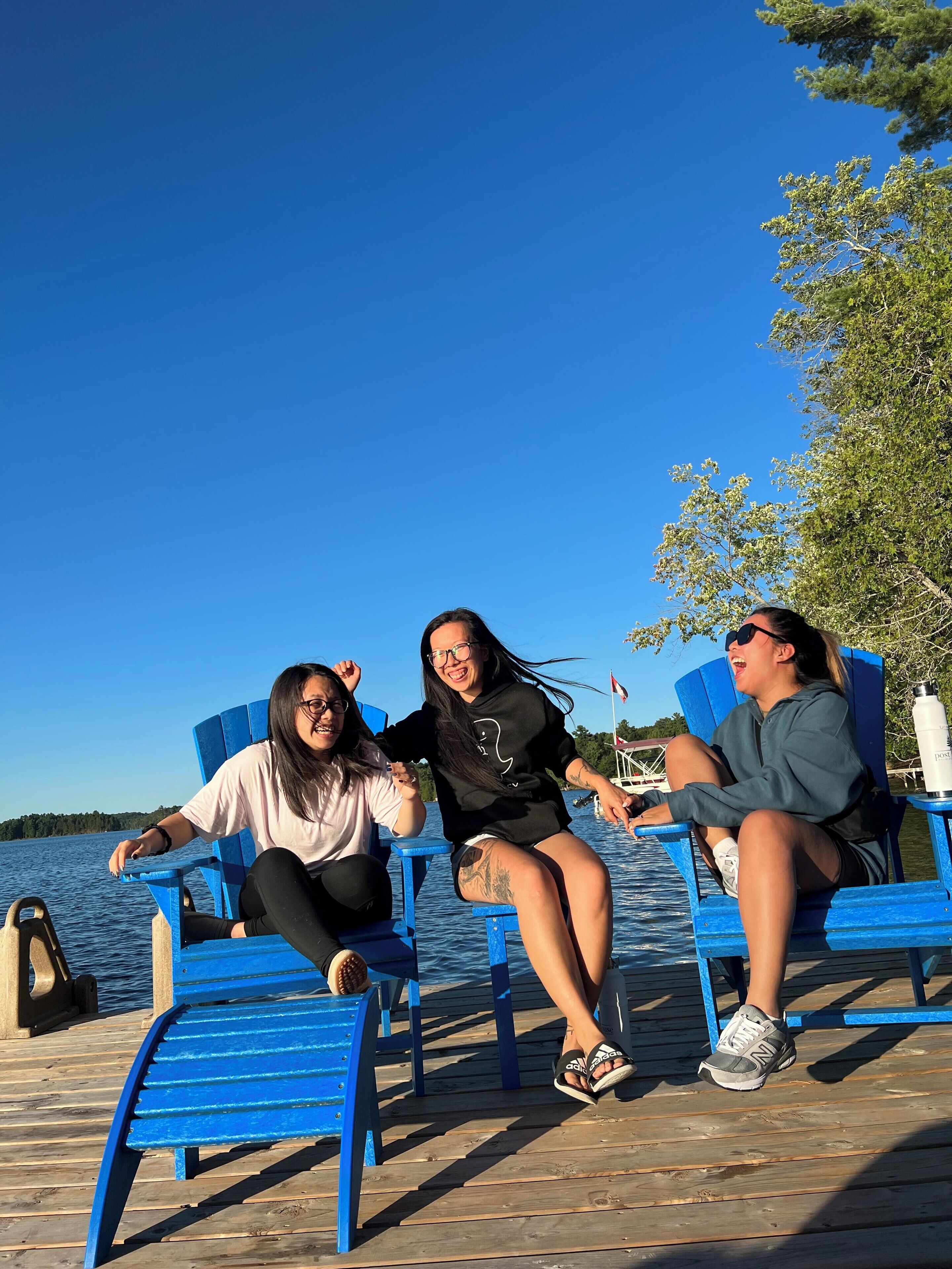 Three people sitting on blue chairs by a wooden dock with a body of water and trees in the background.