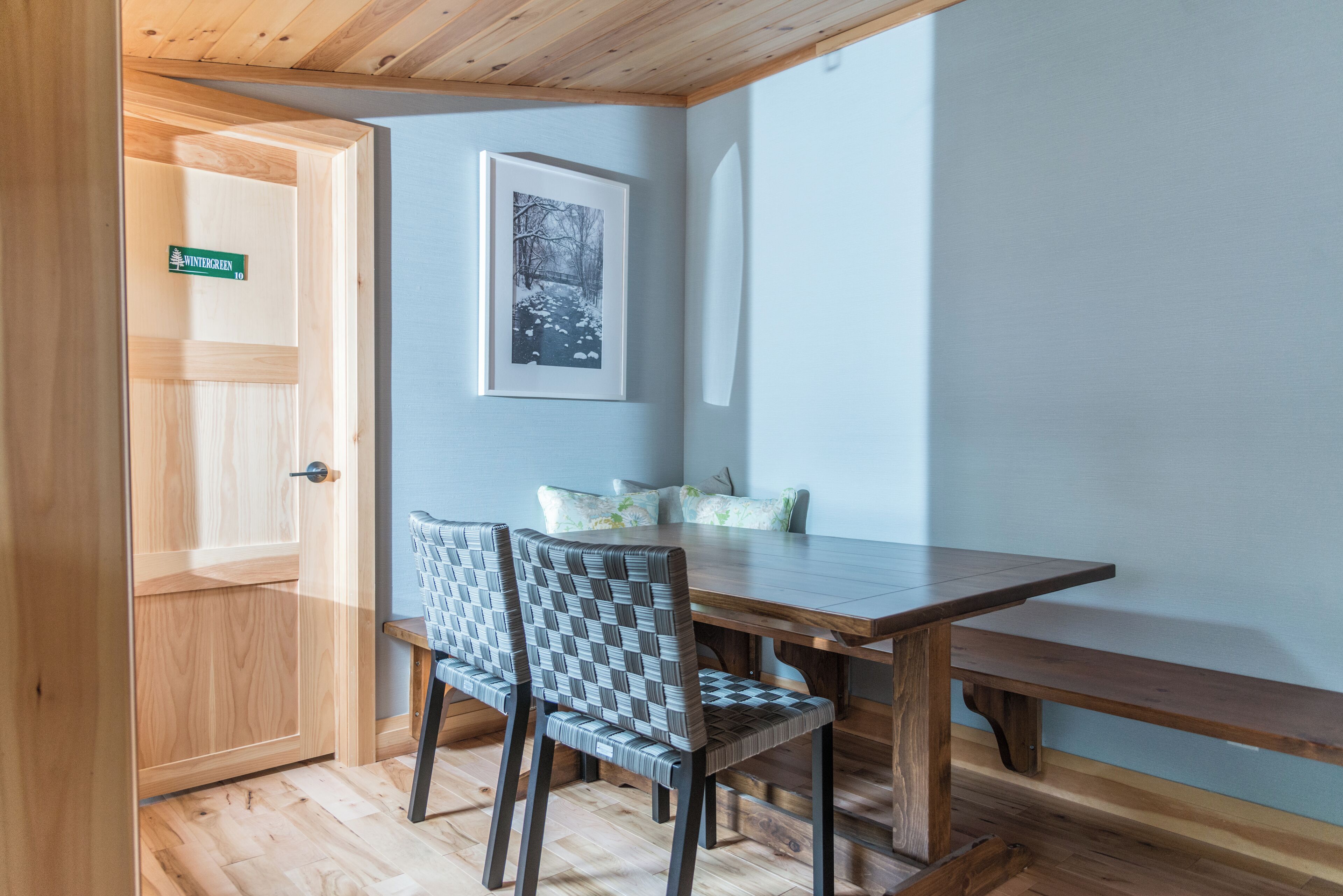 A wooden dining table with two chairs, a framed picture on the wall, and a door with a sign.