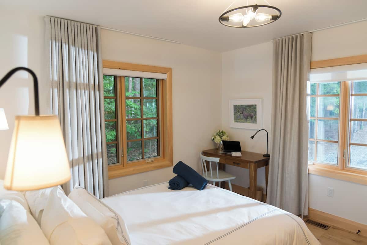 A bedroom with a bed, a desk, and a chair. There is a window with curtains, a lamp, and a picture on the wall.