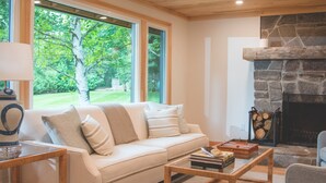 A cozy living room with a beige sofa, a coffee table, and a stone fireplace. Large windows offer a view of greenery outside. - Haliburton Post House (Minden)