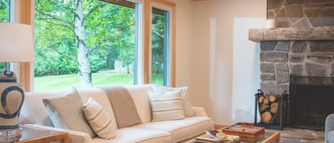 Una sala de estar acogedora con un sofá color beige, una mesa de centro y una chimenea de piedra. Amplias ventanas ofrecen vistas al exterior, con vegetación.