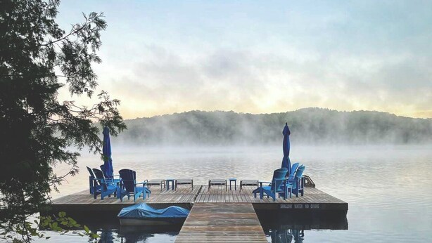 A wooden dock extending into calm water with two blue chairs and umbrellas on each side. - Haliburton Post House (Minden)