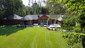 A log cabin with a patio area, surrounded by a well-maintained lawn and dense forest. - Haliburton Post House (Minden)