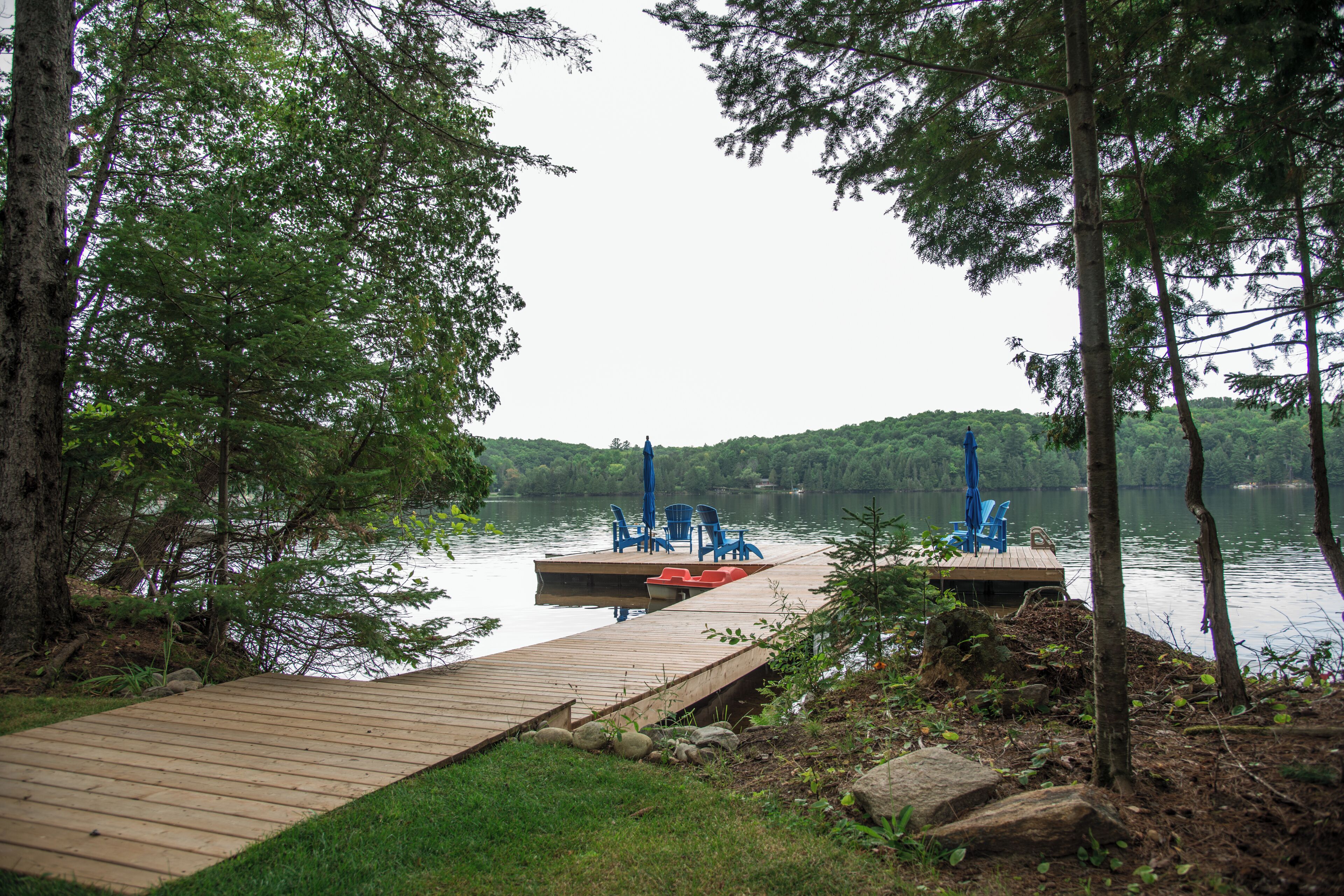 A wooden dock leading to a small dock with blue chairs and a table, surrounded by trees and a lake.