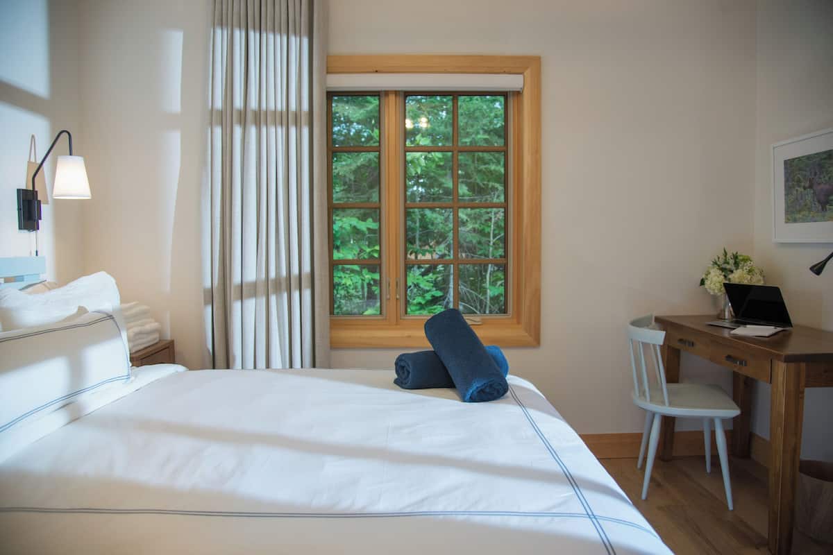 A neatly made bed with white linens, a wooden desk with a laptop, and a window with a view of trees.
