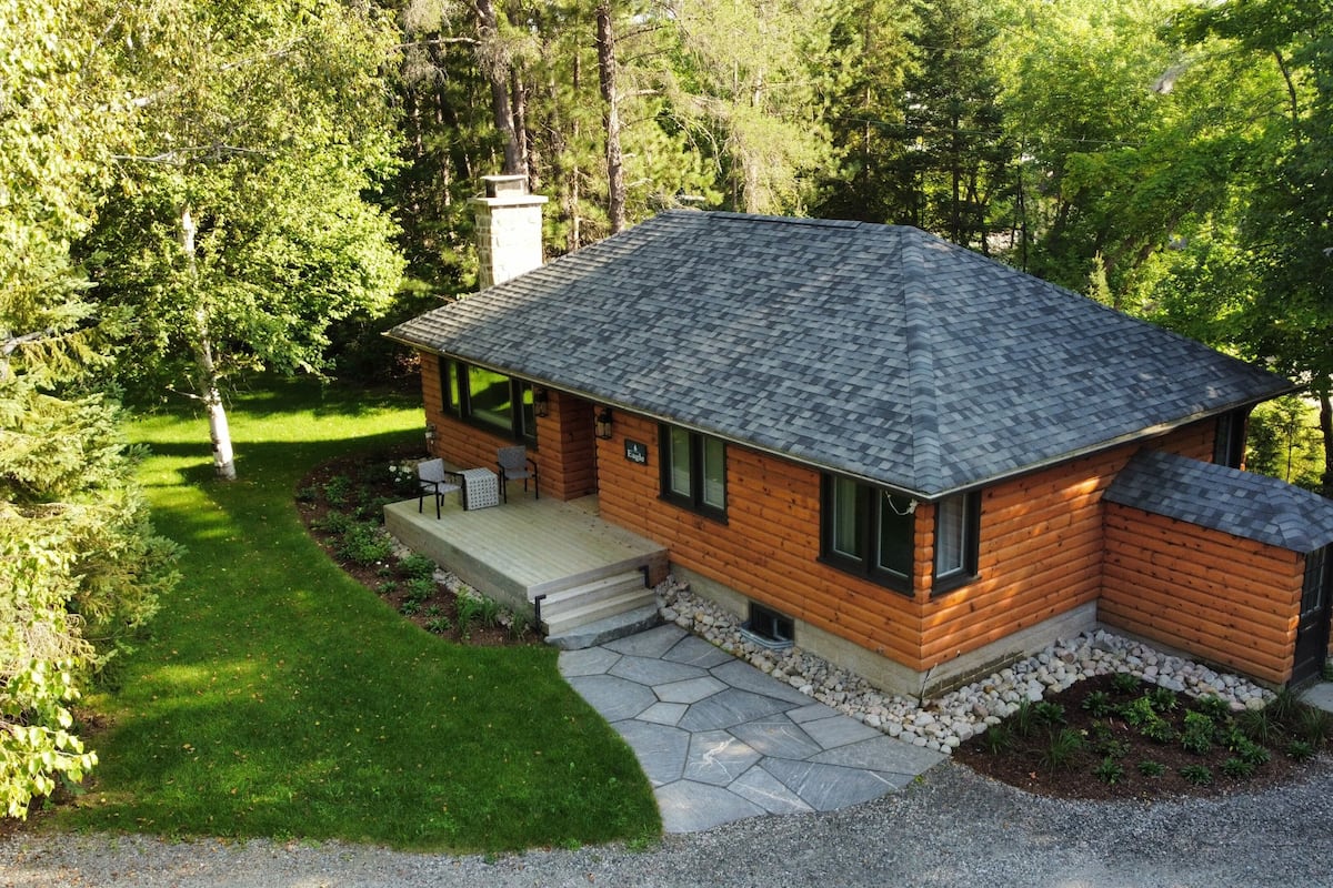 A log cabin with a stone deck, a chimney, and a porch with chairs.