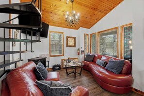 A spacious living room with a red leather sofa, a wooden staircase, and a chandelier. - Stunning 2 Bedroom Cabin In The Woods, Covered Porch, Jacuzzi, Kitchen & Hot Tub (Eureka Springs)
