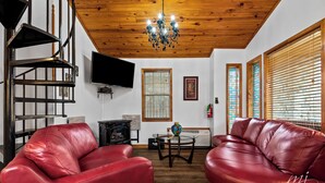 A living room with a red leather sofa, a wooden ceiling, a chandelier, a fireplace, and a television. - Stunning 2 Bedroom Cabin In The Woods, Covered Porch, Jacuzzi, Kitchen & Hot Tub (Eureka Springs)