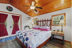 A bedroom with a wooden ceiling, a bed with a floral bedspread, a nightstand with a lamp, and a window with a view of trees. - Stunning 2 Bedroom Cabin In The Woods, Covered Porch, Jacuzzi, Kitchen & Hot Tub (Eureka Springs)