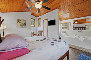 A bedroom with a bed, a ceiling fan, a painting, a television, and a bathroom with a bathtub and sink. - Stunning 2 Bedroom Cabin In The Woods, Covered Porch, Jacuzzi, Kitchen & Hot Tub (Eureka Springs)