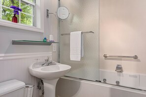 Une salle de bain avec un lavabo blanc, une cabine de douche en verre, une serviette blanche et une fenêtre ornée d’un vase de fleurs violettes.