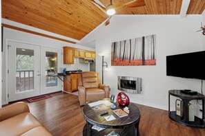 A modern living room with a leather sofa, a round coffee table with magazines, a red decorative item, and a flat-screen TV mounted on the wall. - 1 Bedroom Cabin In The Woods, Covered Porch, Jacuzzi, Kitchen & Hot Tub. (Eureka Springs)