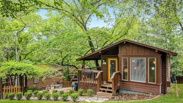 A wooden cabin with a porch, surrounded by lush greenery and trees. - 1 Bedroom Cabin In The Woods, Covered Porch, Jacuzzi, Kitchen & Hot Tub. (Eureka Springs)