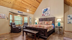 Une chambre avec un plafond en bois, un grand lit avec une literie à motifs floraux, deux chaises, un banc et une petite table avec des lampes.