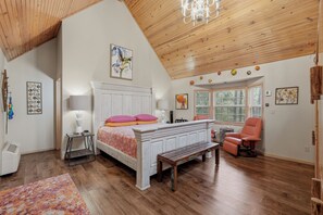 Un dormitorio con techo de madera, cama con ropa de cama rosa y naranja, un banco, una silla y una ventana con persianas.
