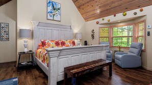 A bedroom with a bed, bedside lamps, a bench, a chair, a window with blinds, and a ceiling with wooden beams. - 1 Bedroom Cabin In The Woods, Covered Porch, Jacuzzi, Kitchenette and Hot Tub. (Eureka Springs)