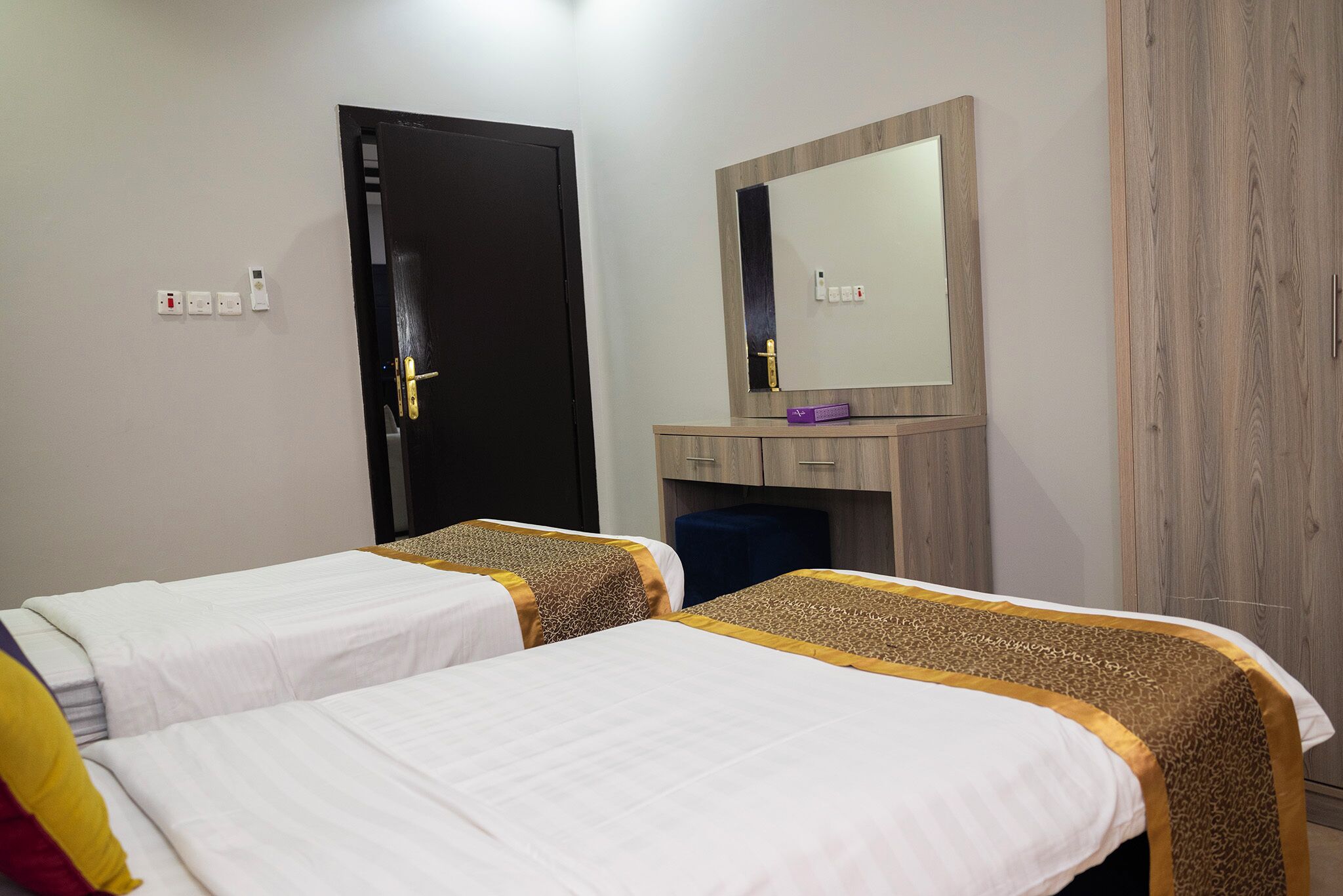 A hotel room with two beds, a wooden wardrobe, a mirror, and a desk with a purple box.