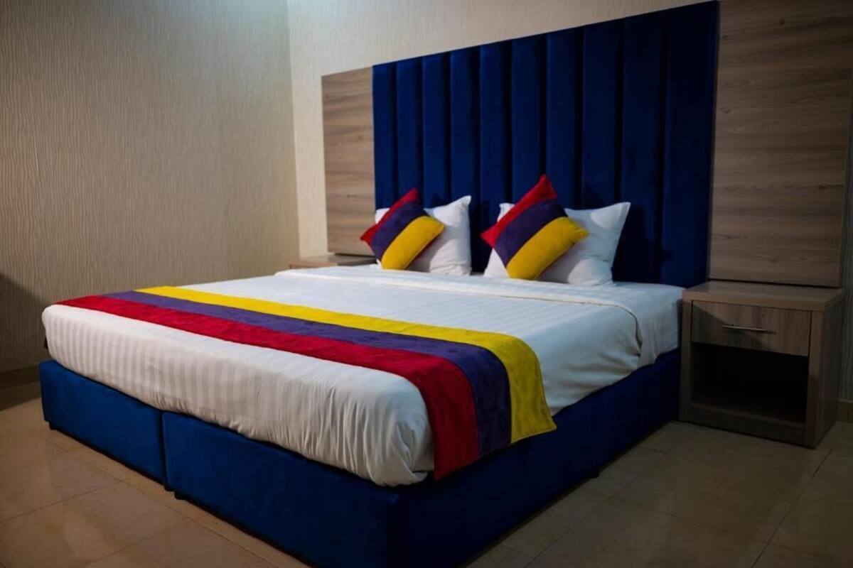 A neatly made bed with a colorful bedspread and pillows, a wooden headboard, and a small bedside table with a drawer.