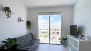 Un salón moderno con un sofá gris, un televisor sobre un mueble blanco y un balcón con vistas a la ciudad.
