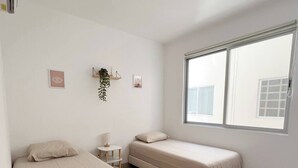 Un dormitorio con dos camas individuales, una mesita pequeña, un estante en la pared con plantas y un ventanal grande.