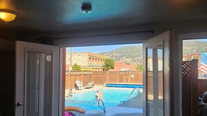 A room with a sliding glass door leading to a backyard with a swimming pool, a barbecue area, and a view of a distant building. - Camaray Motel (Oroville)