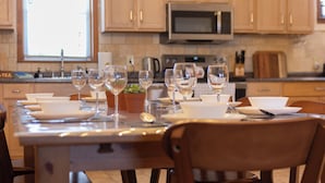 Una mesa de comedor dispuesta con varias copas de vino y platos, una cafetera y un microondas en una cocina con armarios de madera.