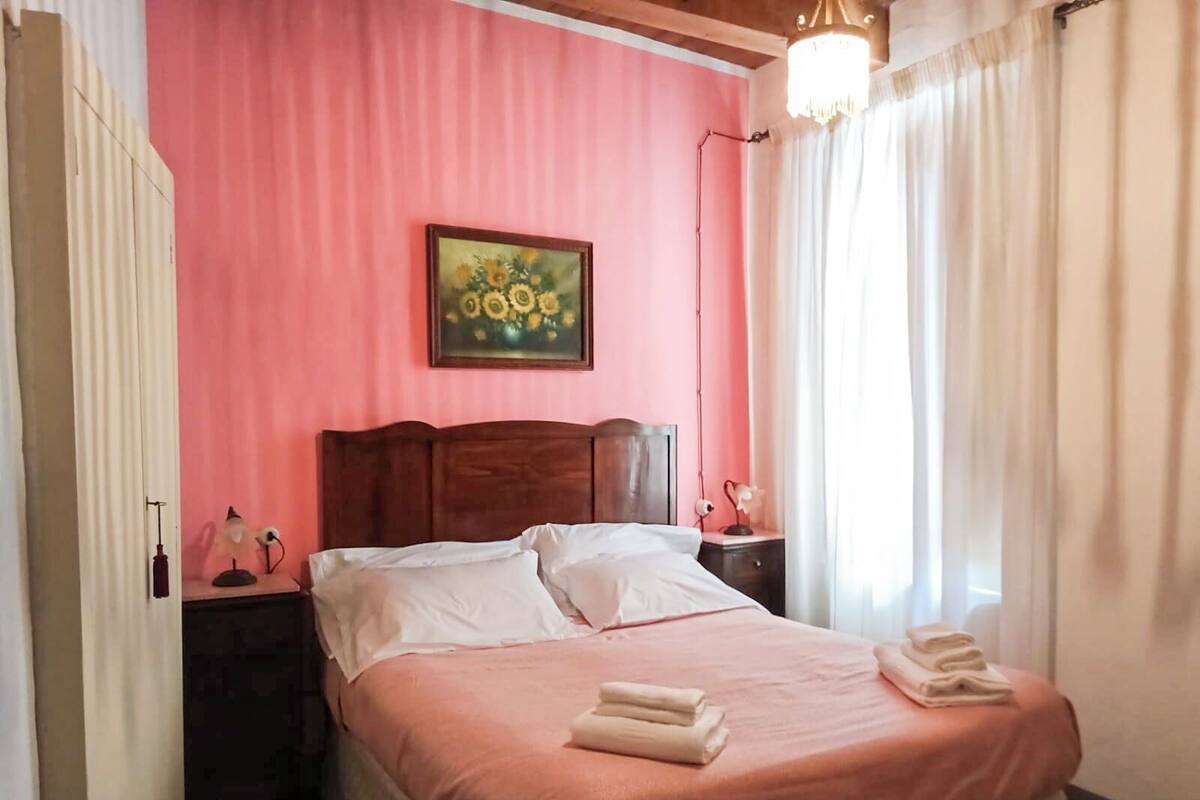 A bedroom with a pink wall, a wooden headboard, a bed with white linens, two bedside tables with lamps, and a painting above the bed.