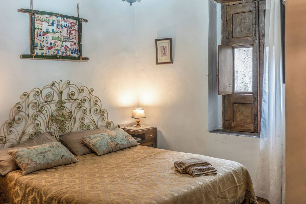 A bedroom with a bed, pillows, a nightstand with a lamp, a wall hanging, and a window.